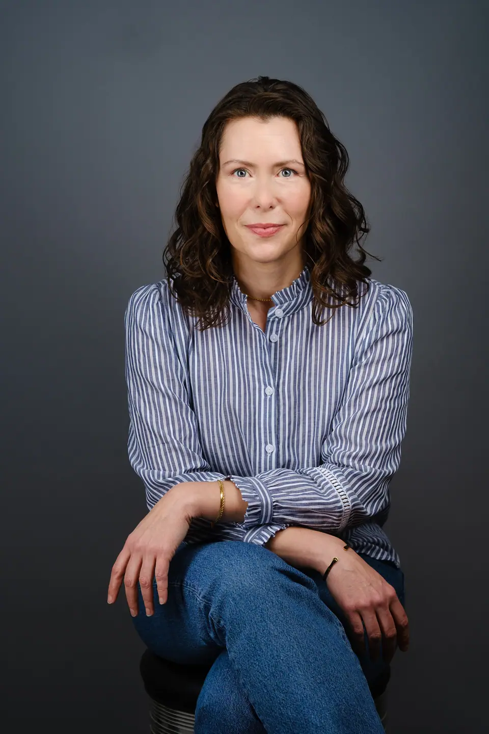 Seated actor portrait of a woman with dark curly hair in a striped shirt and jeans