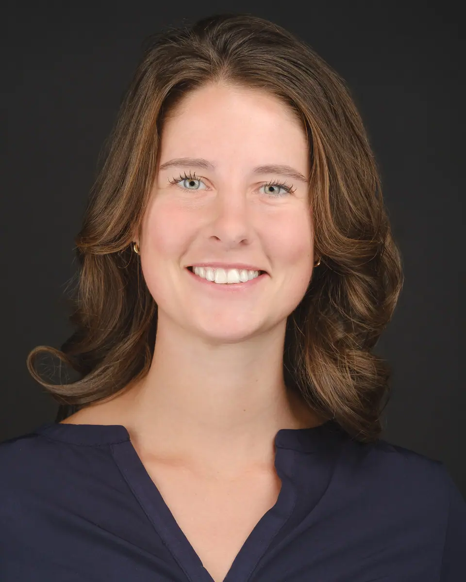 Conference headshot of a smiling woman with wavy brown hair on a black backdrop