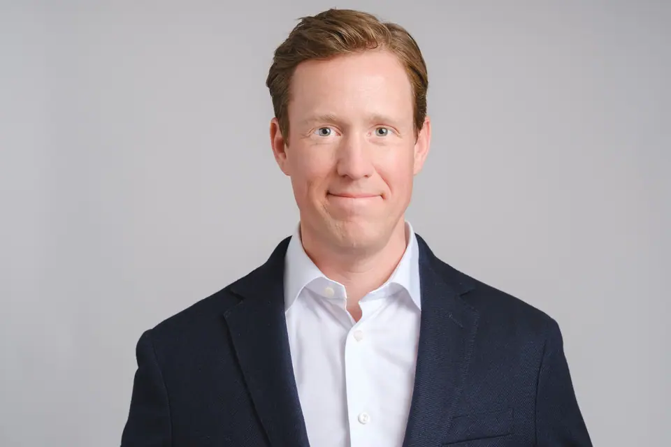 Corporate headshot of a smiling man in a navy blazer on a light grey backdrop