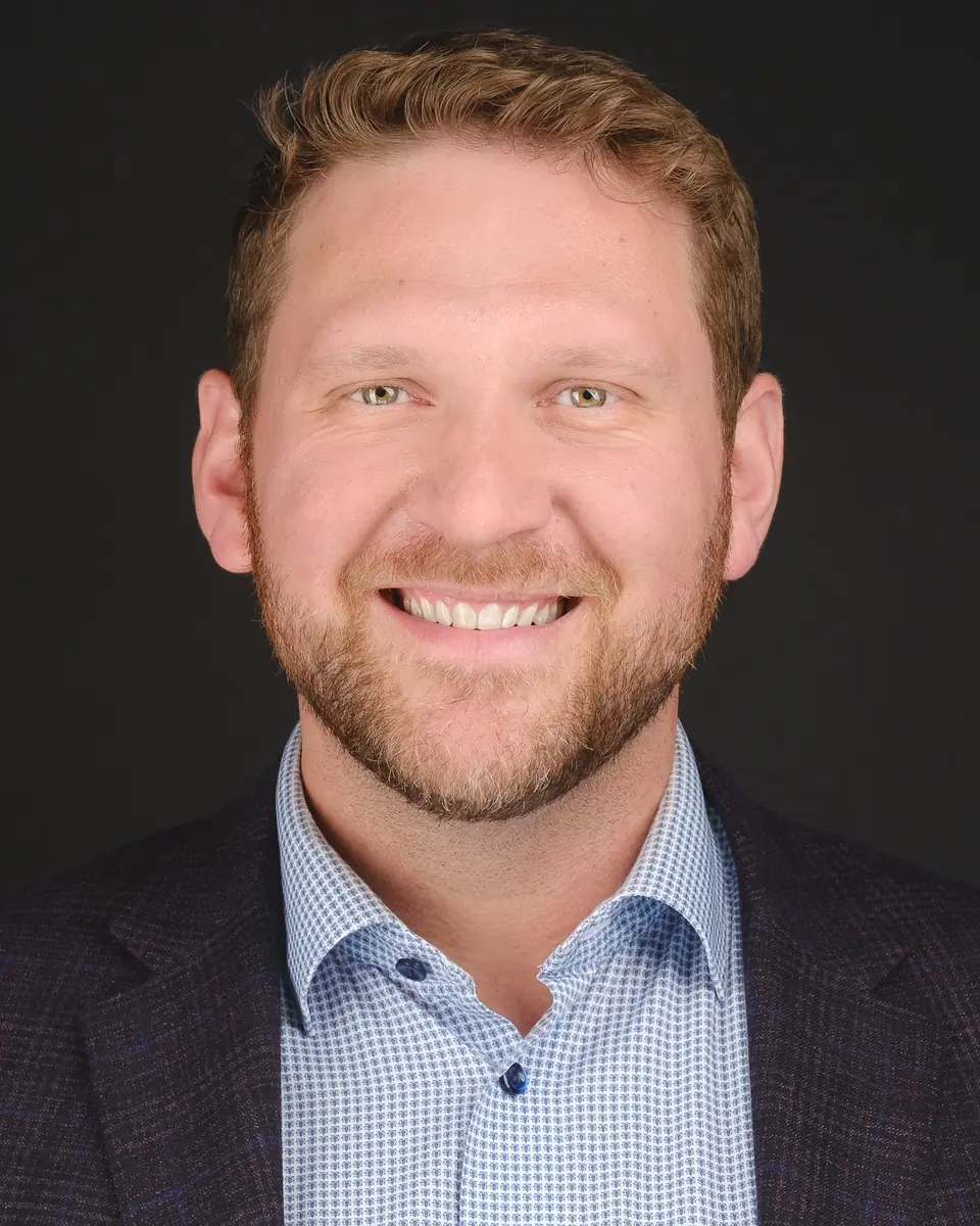 Headshot of a smiling man with a beard in a blazer and checked shirt