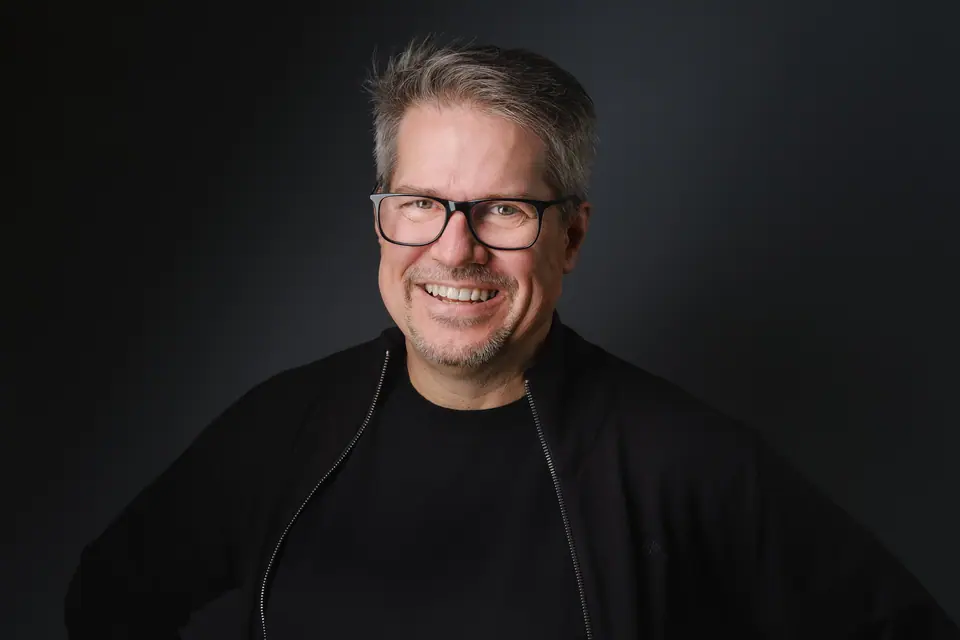 Headshot of a laughing man with glasses in a black jacket on a dark backdrop