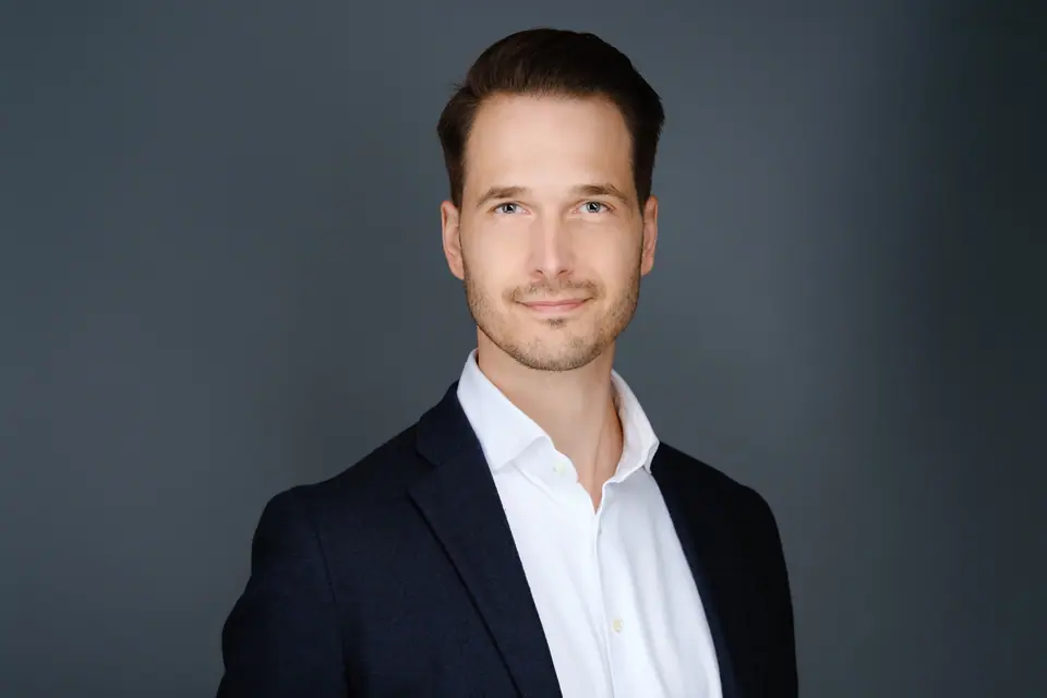 Studio headshot of a smiling man in a navy blazer and open white collar on a grey backdrop