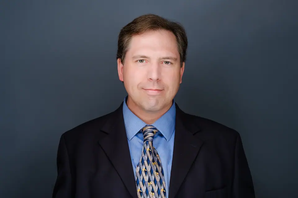 Corporate headshot of a man in a dark suit, blue shirt and patterned tie