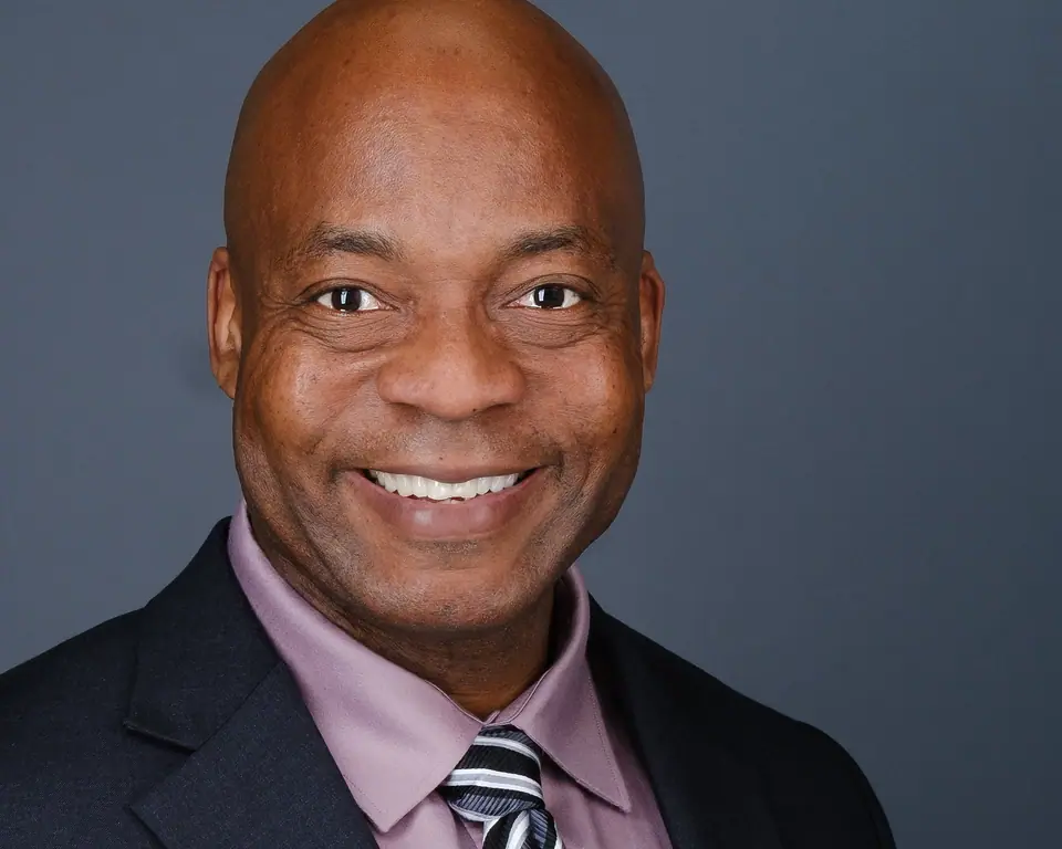 Corporate headshot of a smiling man in a dark suit, pink shirt and striped tie