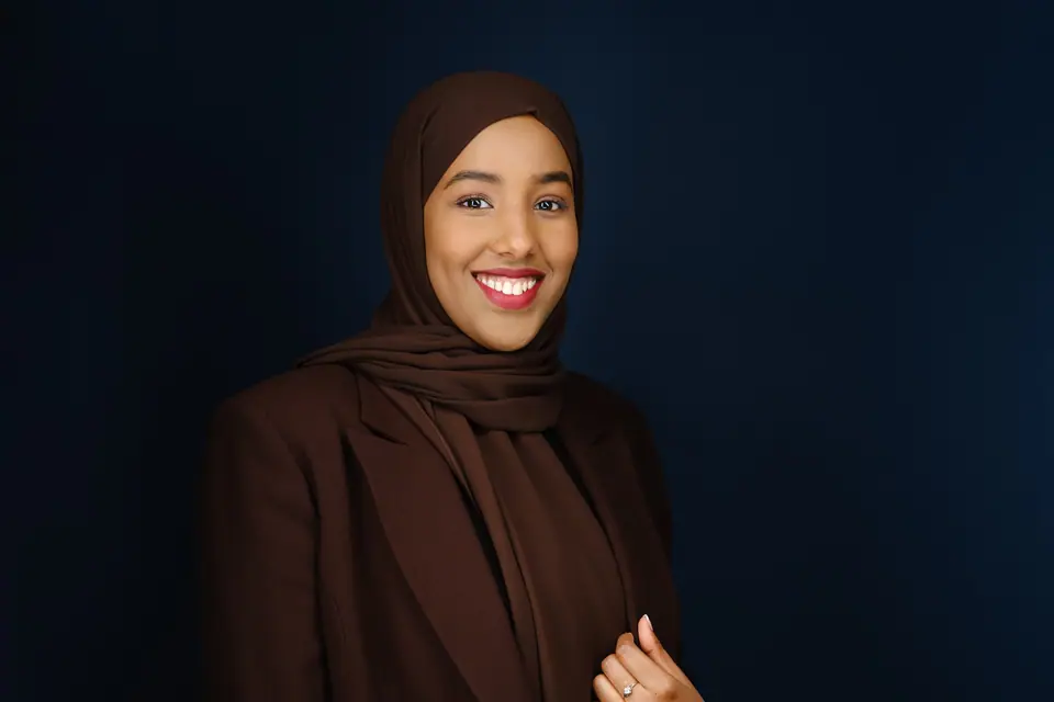 Studio headshot of a smiling woman in a brown hijab and blazer on a deep blue backdrop