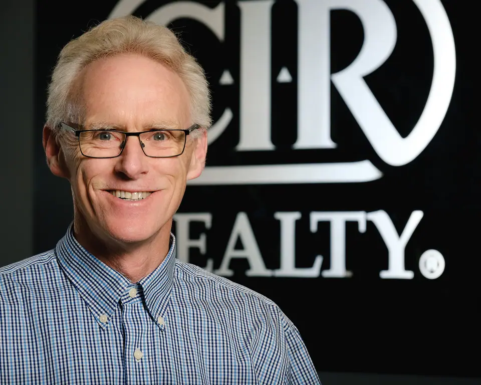 Realtor headshot of a smiling man with glasses in front of his brokerage sign